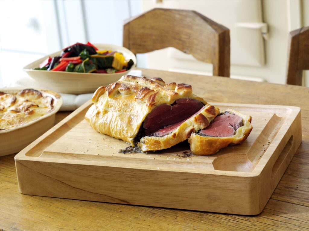 Beef Wellington sits on a wooden cutting board on a dining room table